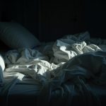 A shadowy, slightly chaotic bedroom with tangled bedding and pillows suggests the challenge of bedtime routines for families.