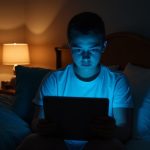 Young adult using a tablet in a softly lit bedroom, illuminated by both a screen’s blue light and a warm bedside lamp, highlighting contrasts between device use and restful ambiance.