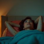 An adult lying awake in bed at night, gazing at the ceiling with thoughtful eyes, surrounded by warm lighting and soft, textured bedding.