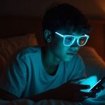 Young adult wearing blue light glasses in bed at night, thoughtfully using a smartphone with warm ambient lighting and cool phone glow.